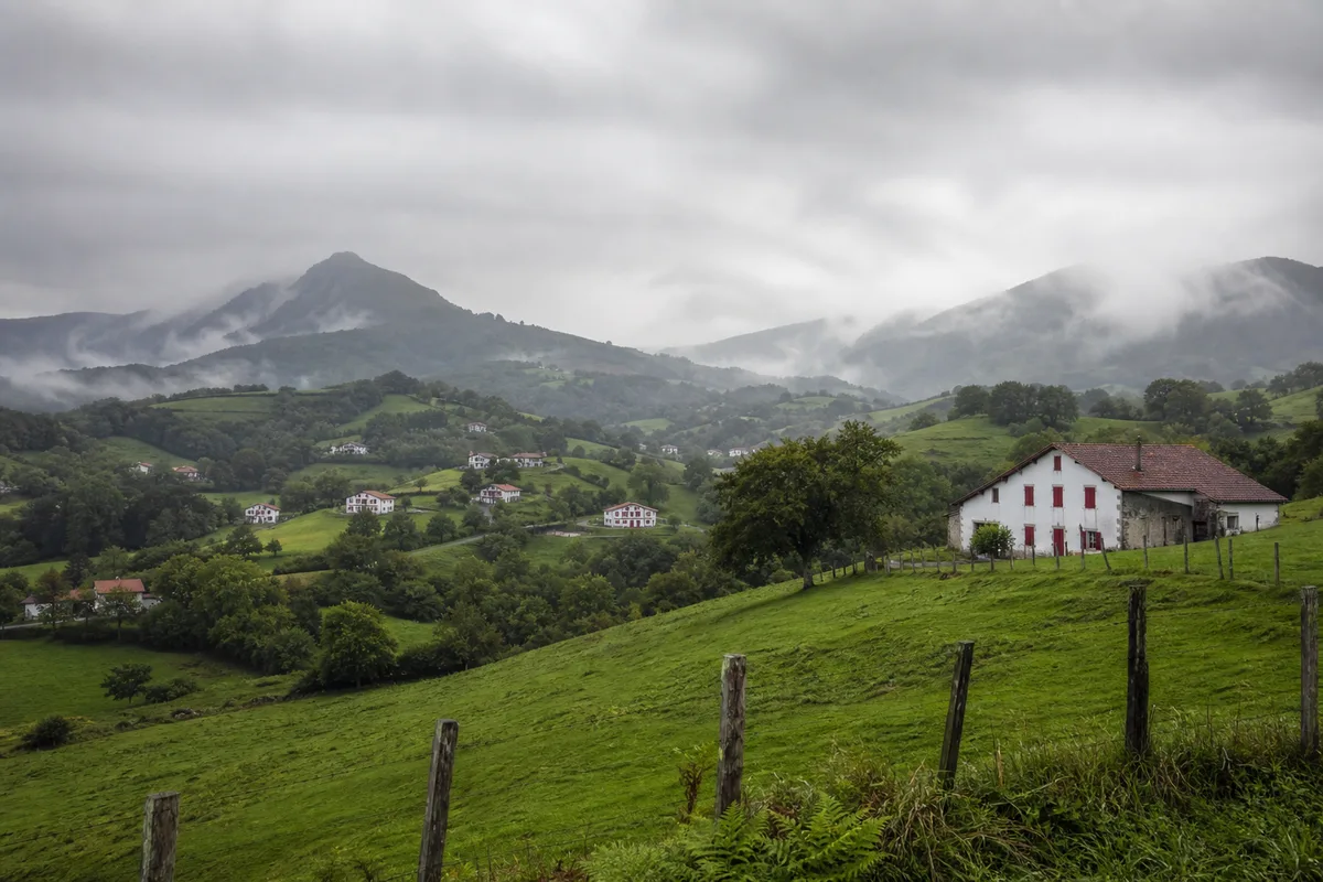 Vallée verdoyante du Pays basque intérieur sous un ciel chargé, maisons traditionnelles aux volets rouges dispersées sur les collines