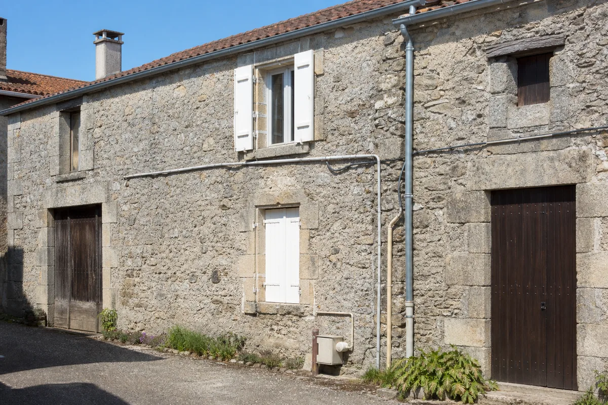 Vue d&rsquo;une maison ancienne en pierre sous un ciel clair, avec un réseau de canalisations extérieures visible le long d&rsquo;un mur